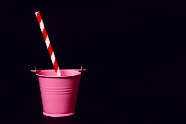 Um balde de brinquedo rosa contra um fundo preto com uma palha de bebida vermelha e branca listrada. O conceito de uma festa ou a necessidade de água em um dia quente. Espaço livre para texto - Foto, Imagem