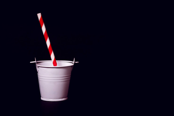 Um balde de brinquedo branco contra um fundo preto com uma palha de bebida vermelha e branca listrada. Conceito de festa com bebidas não padronizadas ou a necessidade de água em um dia quente - Foto, Imagem