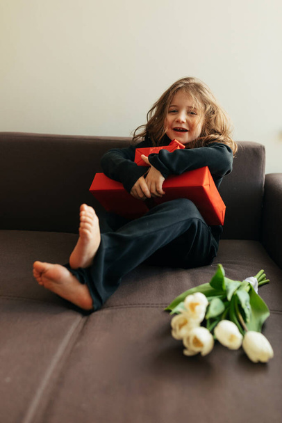 Encantadora niña feliz vistiendo ropa de casa disfrutando de sus regalos y flores en el día de la mujer. Foto de alta calidad - Foto, imagen