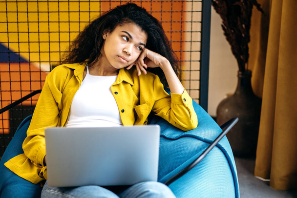 African American young woman sits in cozy armchair, getting bored while studying or working. Beautiful female employee or student looks away, using laptop, thinking about some new ideas - Photo, Image