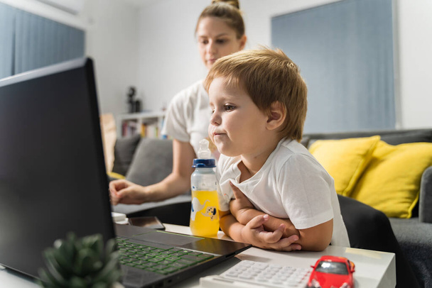 Kleine jongen staan in de voorkant van de computer laptop thuis met zijn moeder in de achtergrond op zoek video besteden tijd online - internet technologie en opgroeien ontwikkelingsconcept - Foto, afbeelding