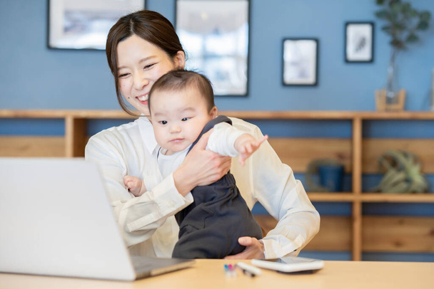 Жінка - азіатка, яка тримає дитину і використовує ноутбук у приміщенні. - Фото, зображення