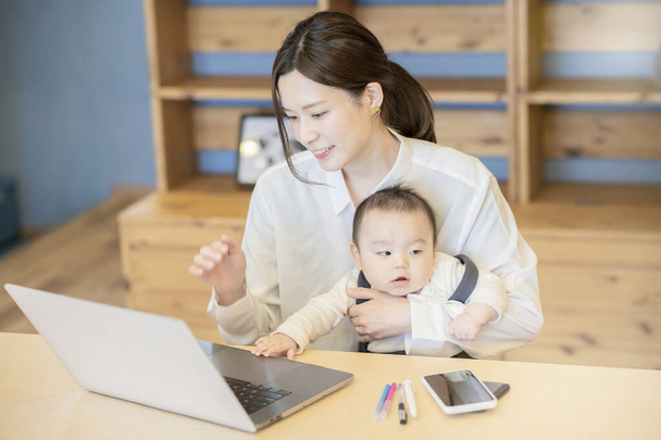 Asian woman holding a baby and operating a laptop indoors - Photo, Image