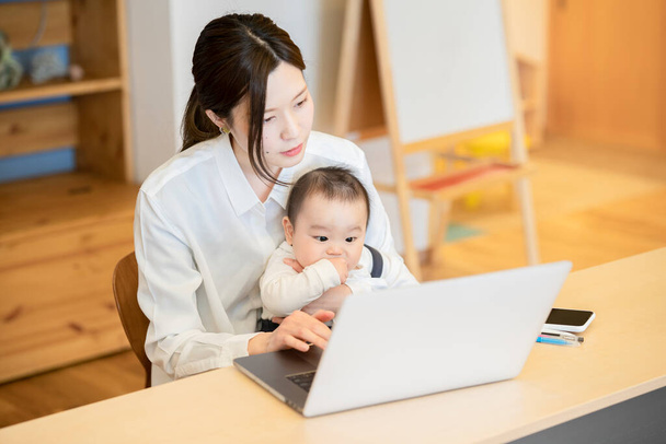 Mulher asiática segurando um bebê e operando um laptop dentro de casa - Foto, Imagem
