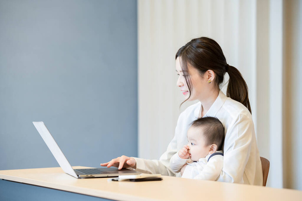 Azjatka trzyma dziecko i operuje laptopa w pomieszczeniu - Zdjęcie, obraz