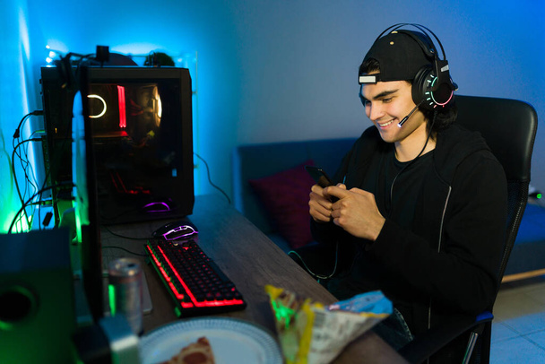 Happy gamer fülhallgatóval mosolyog, miközben sms-ezik és használja a közösségi média az okostelefonján. Latin fiatal férfi fülhallgatóval ül előtt egy játék PC - Fotó, kép