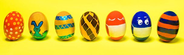 Sarı arka planda çok renkli Paskalya yumurtaları. Yüzü olan yumurtalar karantina sırasında koronavirüse karşı koruyucu maskeler taktılar.. - Fotoğraf, Görsel
