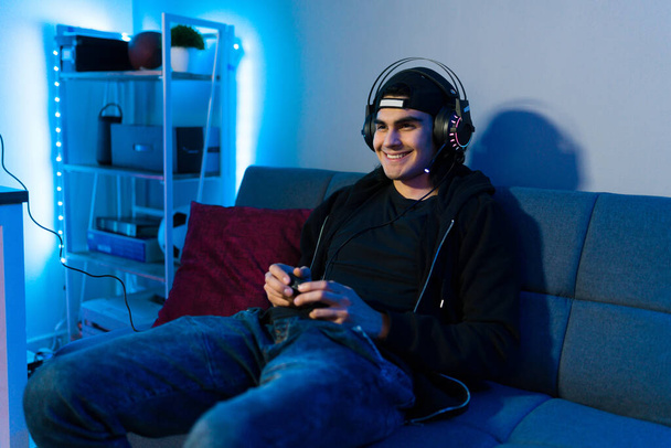 Onnellinen nuori mies, jolla on kuulokkeet sohvalla ja kaukosäädin. Hymyilevä pelaaja pelaa videopeliä ja pitää hauskaa  - Valokuva, kuva
