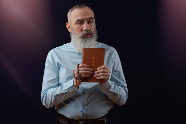 Senior férfi szürke szakállal, világos pólóban, sötét alkalmi kabát kezében egy könyvet Szent Biblia fa rózsafüzér egy kereszt ellen sötét háttér lila kiemeli - Fotó, kép