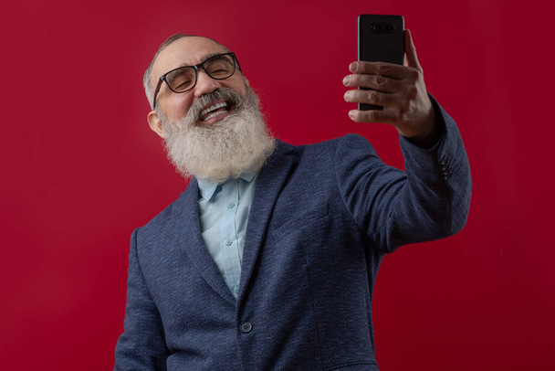 Idősebb boldog férfi, szürke szakállal, világos pólóban, sötét alkalmi kabátban, kimenő néz egy okostelefonba, csinál egy szelfit vagy csevegik. - Fotó, kép