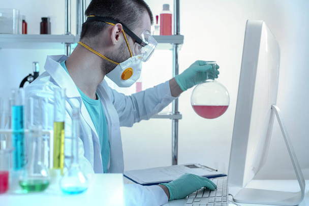 Asystent laboratorium chemicznego w okularach ochronnych i masce bada eksperymentalne probówki pracujące z danymi na komputerze. - Zdjęcie, obraz