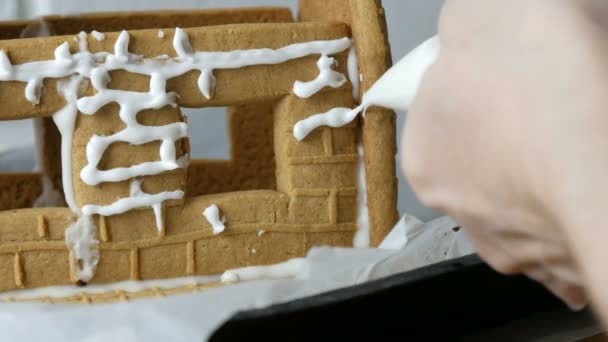 Nő díszíti mézeskalács ház tészta táska fehér cukor édes cukormáz, kezét fehér tégla háttér. Főzés, házi készítésű mézeskalács ház karácsonyi ünnepekre. Újévi hagyományok - Felvétel, videó