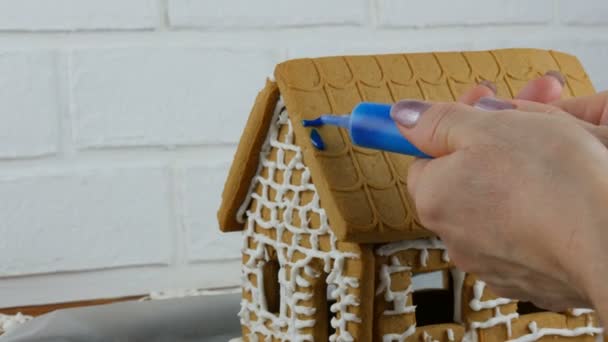 Nő díszíti mézeskalács ház kék édes cukormáz, kezét fehér tégla háttér. Főzés, házi készítésű mézeskalács ház karácsonyi ünnepekre. Újévi hagyományok - Felvétel, videó