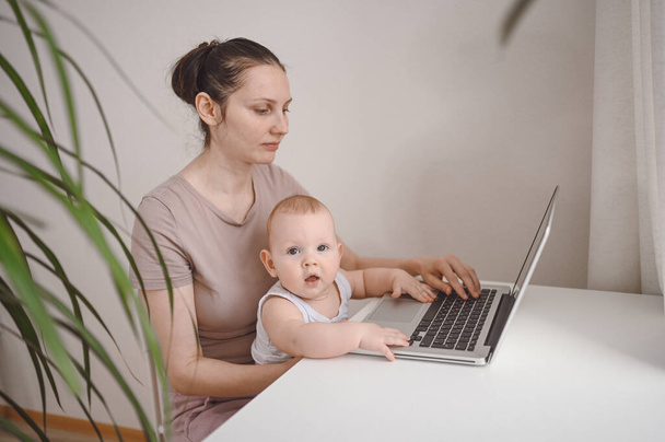 Junge Mütter arbeiten während der Quarantäne von zu Hause aus mit Laptop, kleines süßes Kleinkind auf dem Schoß. Home Office, Elternschaft. Fernarbeit, Fernstudium. Virtuelles Ereignis Videokonferenz - Foto, Bild