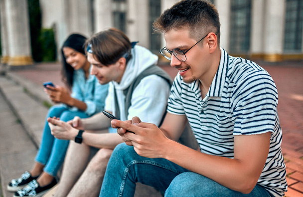 Un grupo de estudiantes se sientan en las escaleras fuera del campus y usan sus teléfonos inteligentes. - Foto, imagen