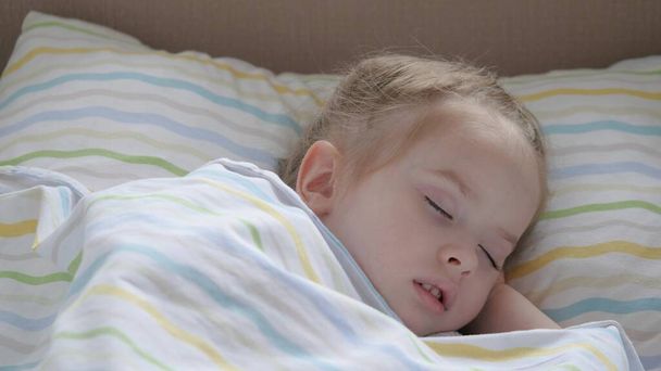 幸せな赤ん坊は赤ん坊のベッドで眠り、笑顔。母さんは疲れている娘を寝かしつけた。かわいい子供が彼のベッドの中の保育園で休んでいる。幸せな家族と子供時代のコンセプト。健康な子供たち. - 写真・画像
