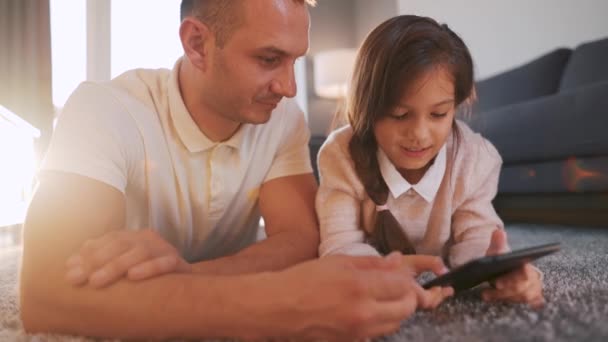 Padre e hija disfrutando del tiempo juntos y utilizando una tableta para el entretenimiento familiar mientras están acostados en el suelo en la sala de estar en casa. - Metraje, vídeo
