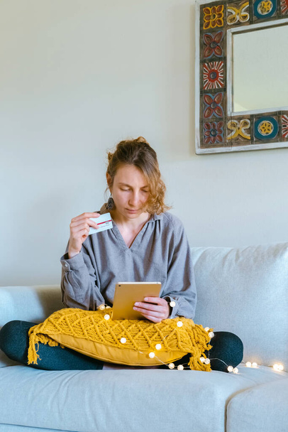 Молода жінка робить онлайн покупки (вибираючи подарунки) сидячи на дивані в затишному домашньому інтер'єрі
. - Фото, зображення