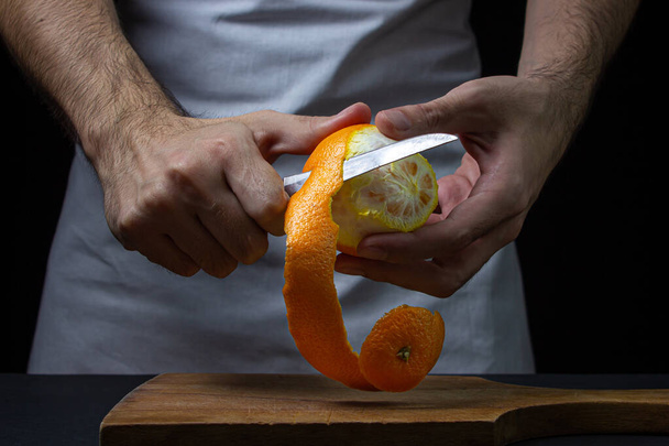 Pomarańczowy na ciemnym tle. Szef kuchni obiera pomarańczę. mężczyzna ręce wyciąć pomarańczowy skórka na czarny tło - Zdjęcie, obraz