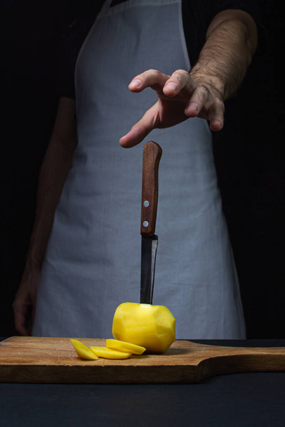 Kartoffeln auf dunklem Hintergrund. Messer in Kartoffeln auf schwarzem Hintergrund. Hand greift zum Messer - Foto, Bild