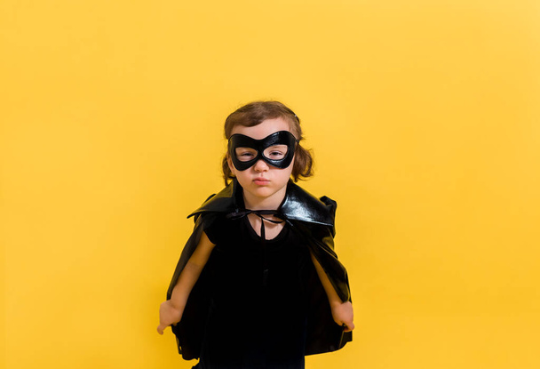 A small supergirl in a black mask and Cape on a yellow isolated background with space for text. - Photo, Image