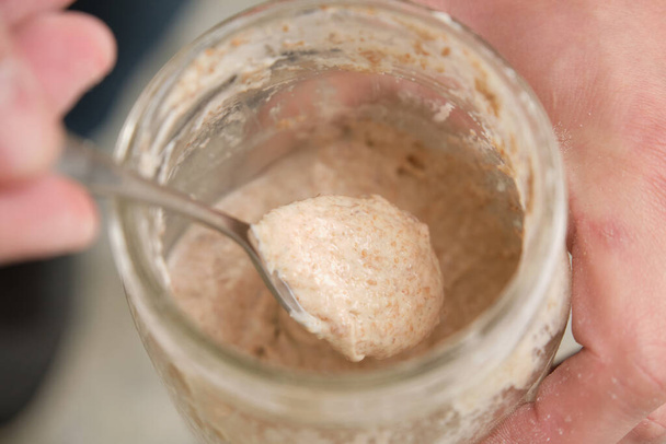 Close up of hands with a spoon grabbing part of sourdough starter. Fermented culture to prepare bread. - Photo, Image