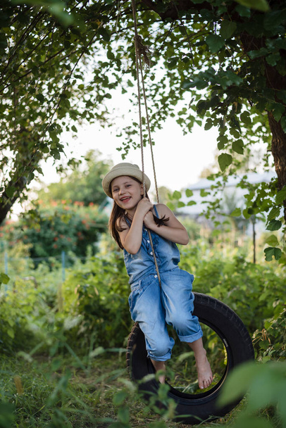 Holčička jezdí na houpačce v zahradě. Dívka má na sobě letní džínovou kombinézu a na hlavě slamák. Letní zábava pro děti - Fotografie, Obrázek