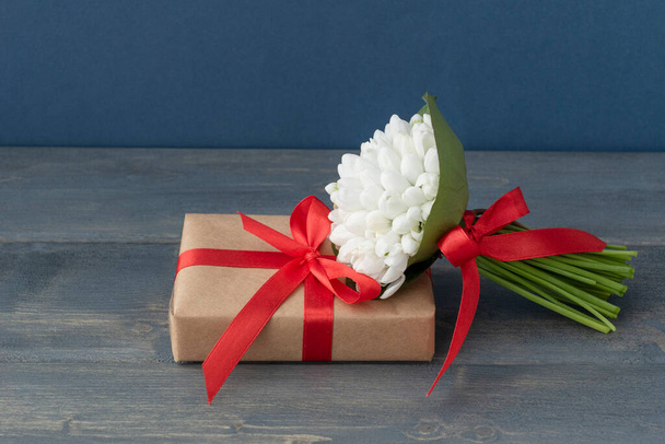 Greeting concept, white snowdrops with red ribbons lie on wrapped giftbox on dark wooden background, lots of copyspace. First spring flowers - Photo, Image