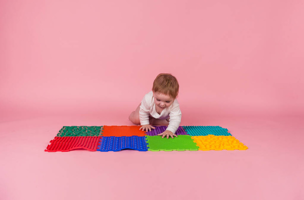 Una niña en un body blanco se arrastra a cuatro patas sobre una alfombra ortopédica sobre un fondo rosa con espacio para el texto - Foto, imagen