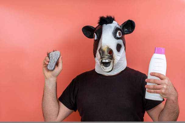 άτομο με μάσκα αγελάδας που δείχνει ένα σφουγγάρι και σαπούνι ντους σε ροζ φόντο - Φωτογραφία, εικόνα