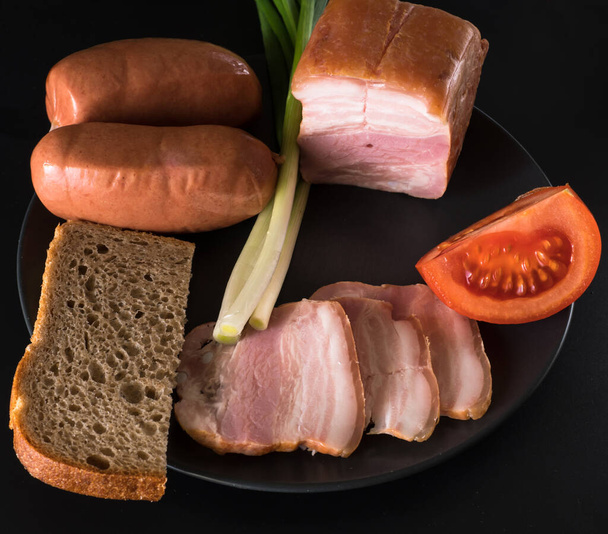 Comida, manteca de cerdo, cebollas verdes, tomate, pan negro, salchichas, en un plato negro sobre un fondo negro - Foto, imagen