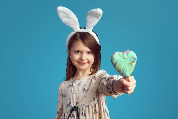 Rozkošná okouzlující dívka drží sušenku ve tvaru srdce, nosí králičí uši přes modrou stěnu - Fotografie, Obrázek