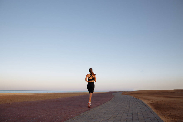 Siyah eşofmanlı ve spor ayakkabılı bir kız gün batımında deniz kenarında koşuyor. - Fotoğraf, Görsel