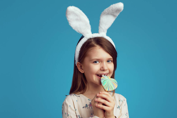 Nadšený holčička v králičí uši kousání srdce ve tvaru sušenky, izolované přes modrou stěnu - Fotografie, Obrázek