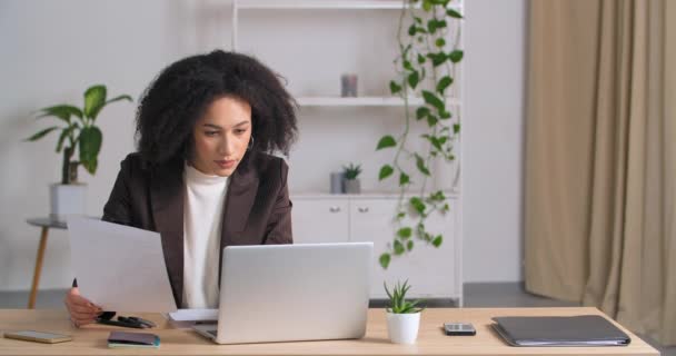 Portrét mladé profesionální afro-americké ženy ve společenském obleku, která sedí u psacího stolu v kanceláři a prohlíží si dokumenty, analyzuje data, píše na notebooku, pracuje během uzamykání - Záběry, video