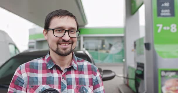 Close-up van een vrolijke knappe man met een bril bij zijn auto terwijl hij tankt bij een benzinestation of diesel. Portret van een knappe jongen. Auto tanken. Zakenman op zoek naar camera - Video