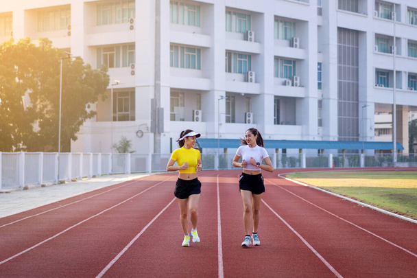 Due giovani donne asiatiche in abiti sportivi che fanno jogging sulla pista da corsa nello stadio cittadino al mattino di sole per mantenere il fitness e uno stile di vita sano. Giovani donne di fitness corrono sulla pista dello stadio. Sport e tempo libero - Foto, immagini