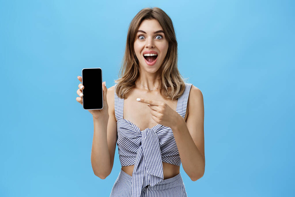 Aufgeregt glückliche Frau mit klappernden Zähnen kaufte schließlich nagelneue Smartphone-Halterung in der Hand zeigt auf Handy-Bildschirm zeigt coole App breit lächeln vor Freude gegen blaue Wand - Foto, Bild
