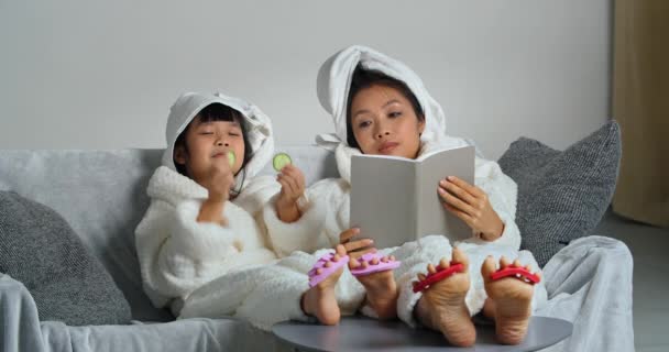 Dos chicas asiáticas de diferentes generaciones en albornoces, madre e hija se relajan después de la ducha en el sofá libro de lectura niño pone círculos frescos de pepino en los ojos hace máscara natural comer verduras - Imágenes, Vídeo