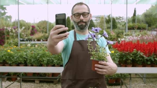 Felice fiorista maschio in possesso di fiore in vaso e prendendo selfie - Filmati, video