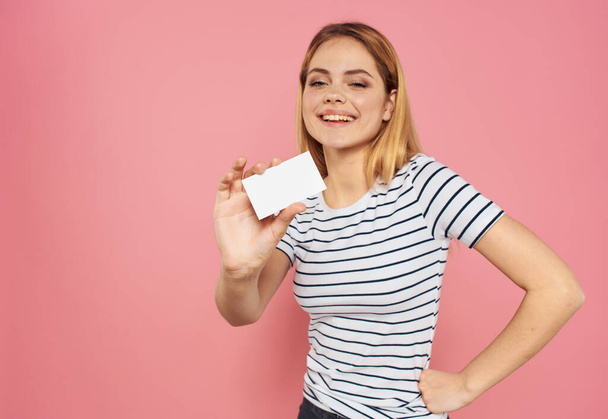 Woman with a business card and in a striped T-shirt on a pink background hand on the waist - Photo, Image