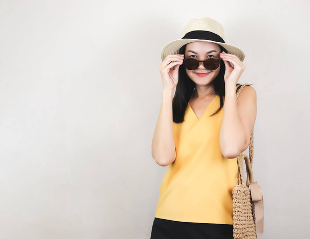Portrét asijské ženy ve žluté košili bez rukávů a klobouku, držící tkanou tašku, stojící na bílém pozadí, sundávající si sluneční brýle, usmívající se a hledící do kamery. Letní koncept. - Fotografie, Obrázek
