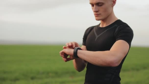 The athlete is including training on his sports watch. He is standing in a field of green grass. The camera is rotating around him. 4K - Footage, Video