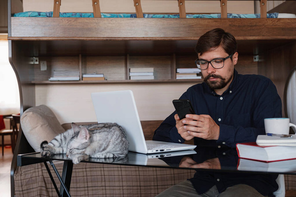 Молодий бородатий бізнесмен сидить в домашньому офісі за столом і використовує ноутбук, поруч сидить сірий кіт. На столі стоїть смартфон, папір, книги, чашка кави. Концепція роботи вдома під час карантину
 - Фото, зображення
