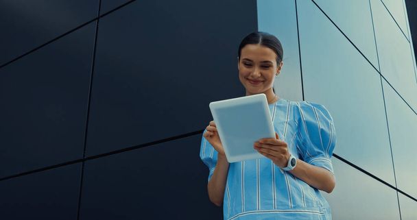 Tiefansicht einer glücklichen Frau in Bluse mit digitalem Tablet, während sie in der Nähe eines Gebäudes steht - Foto, Bild
