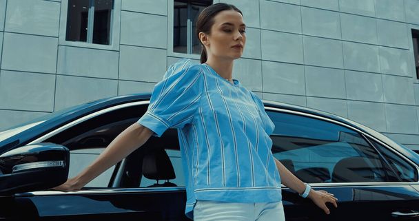young brunette woman in blue blouse posing near black car - Photo, Image