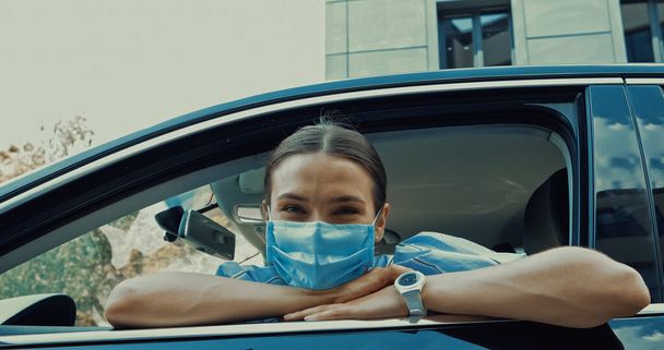 giovane donna in maschera medica seduta sul sedile del conducente e guardando la fotocamera dalla finestra aperta - Foto, immagini