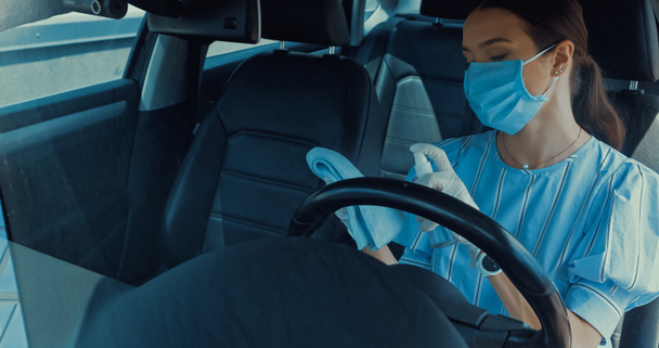 γυναίκα με ιατρική μάσκα ψεκασμού απολυμαντικό σε κουρέλι στο εσωτερικό του αυτοκινήτου - Φωτογραφία, εικόνα