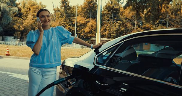 Benzin istasyonunda arabaya yakıt doldururken akıllı telefondan konuşan mutlu bir kadın. - Fotoğraf, Görsel