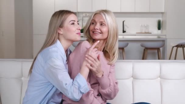 2人の女性、美しい笑顔のシニアの母親と彼女の大人の娘が自宅でソファに座って、一緒に過ごす時間を楽しんで抱き合っています。家族愛の概念. - 映像、動画